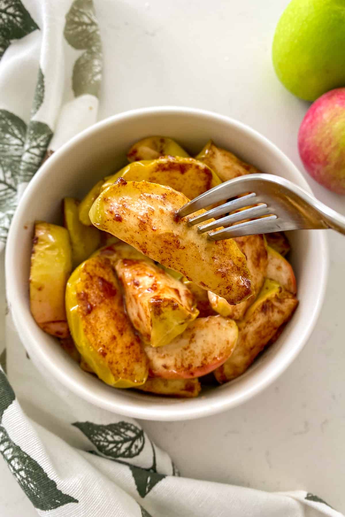 Fork holding up baked apple wedge with bowl of baked apples below.