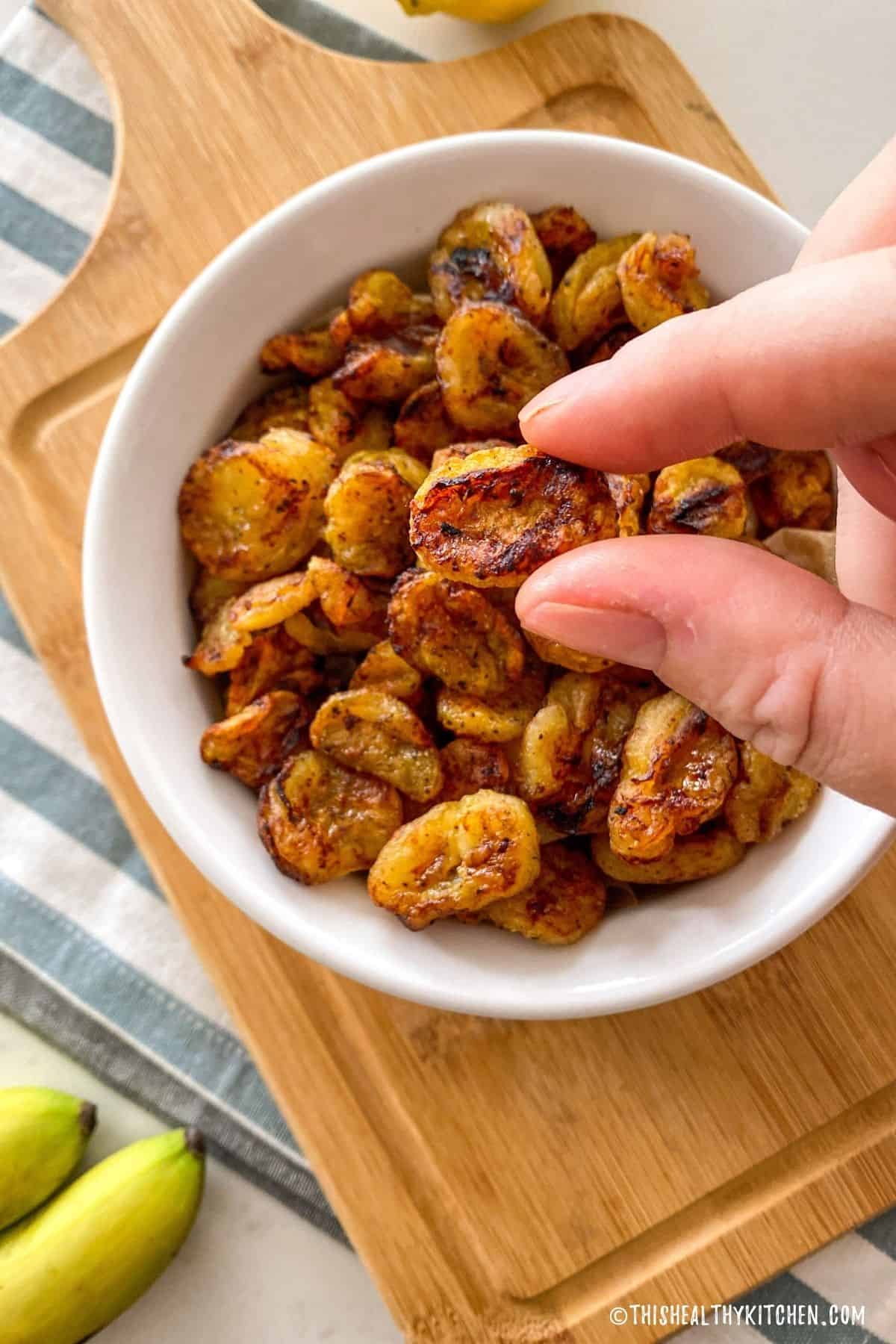 Hand holding up one slice of fried banana above bowl of banana chips.