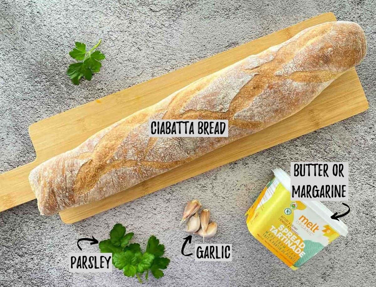 Loaf of bread on cutting board with butter, garlic, and parsley scattered around.