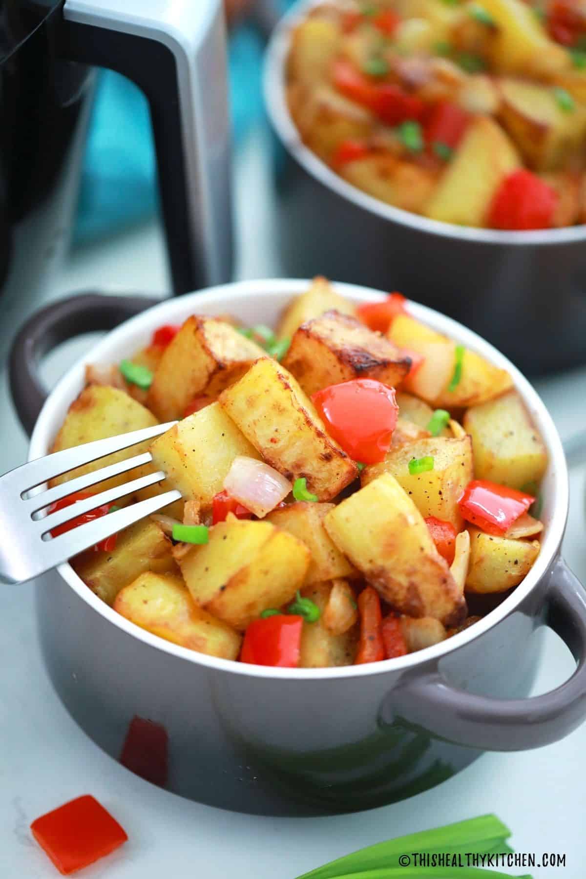 Fork piercing through a breakfast potato above bowl of more breakfast potatoes.