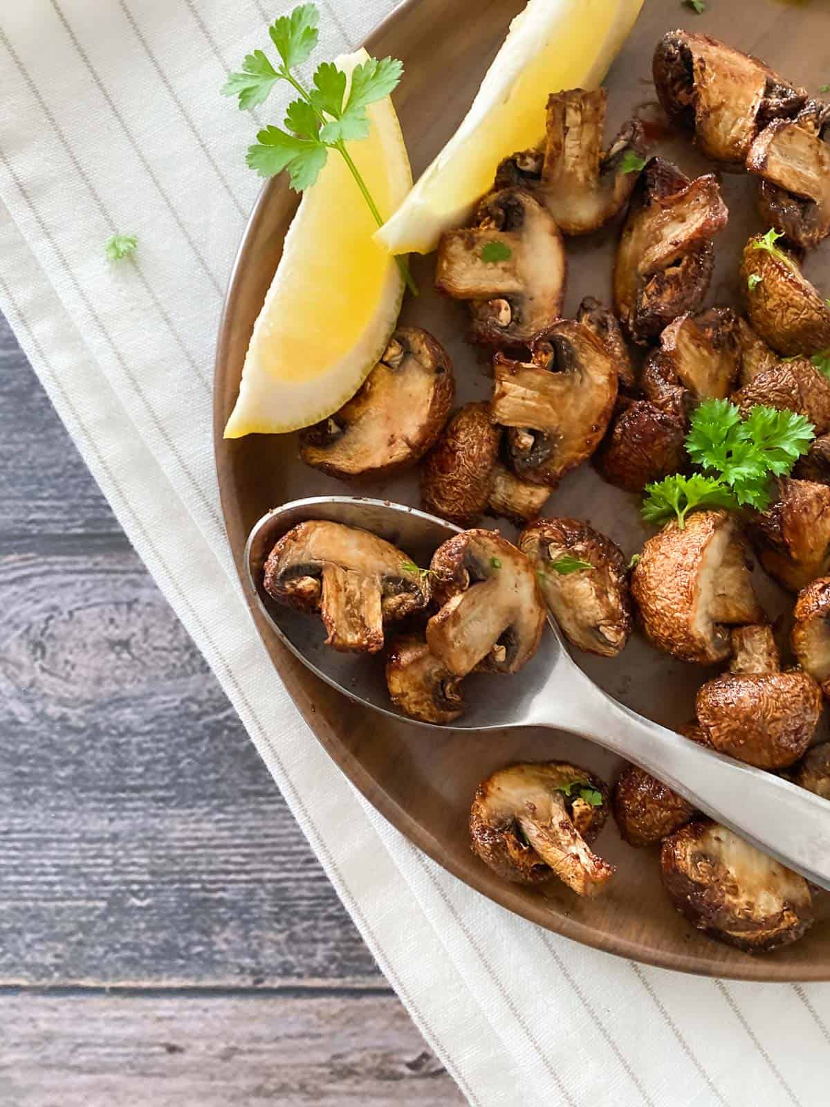 Bamboo plate of mushrooms with spoon of mushrooms resting on top and lemon wedges inside.