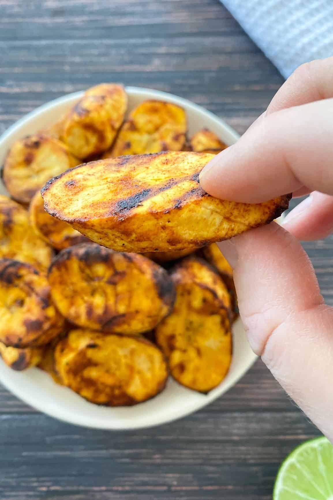 Hand holding up slice of air fried plantain over plate with additional slices.