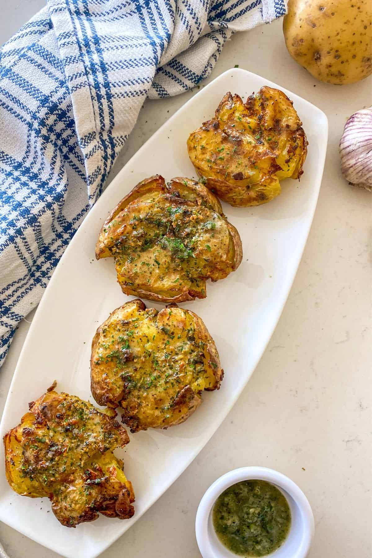 Smashed potatoes on white serving dish with garlic butter in small bowl beside it.