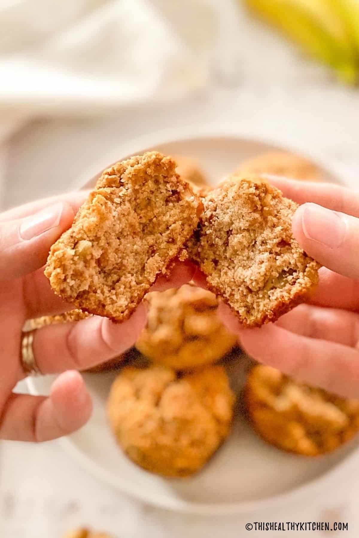Hands holding up a muffin that's split in half with plate of remaining muffins below it.