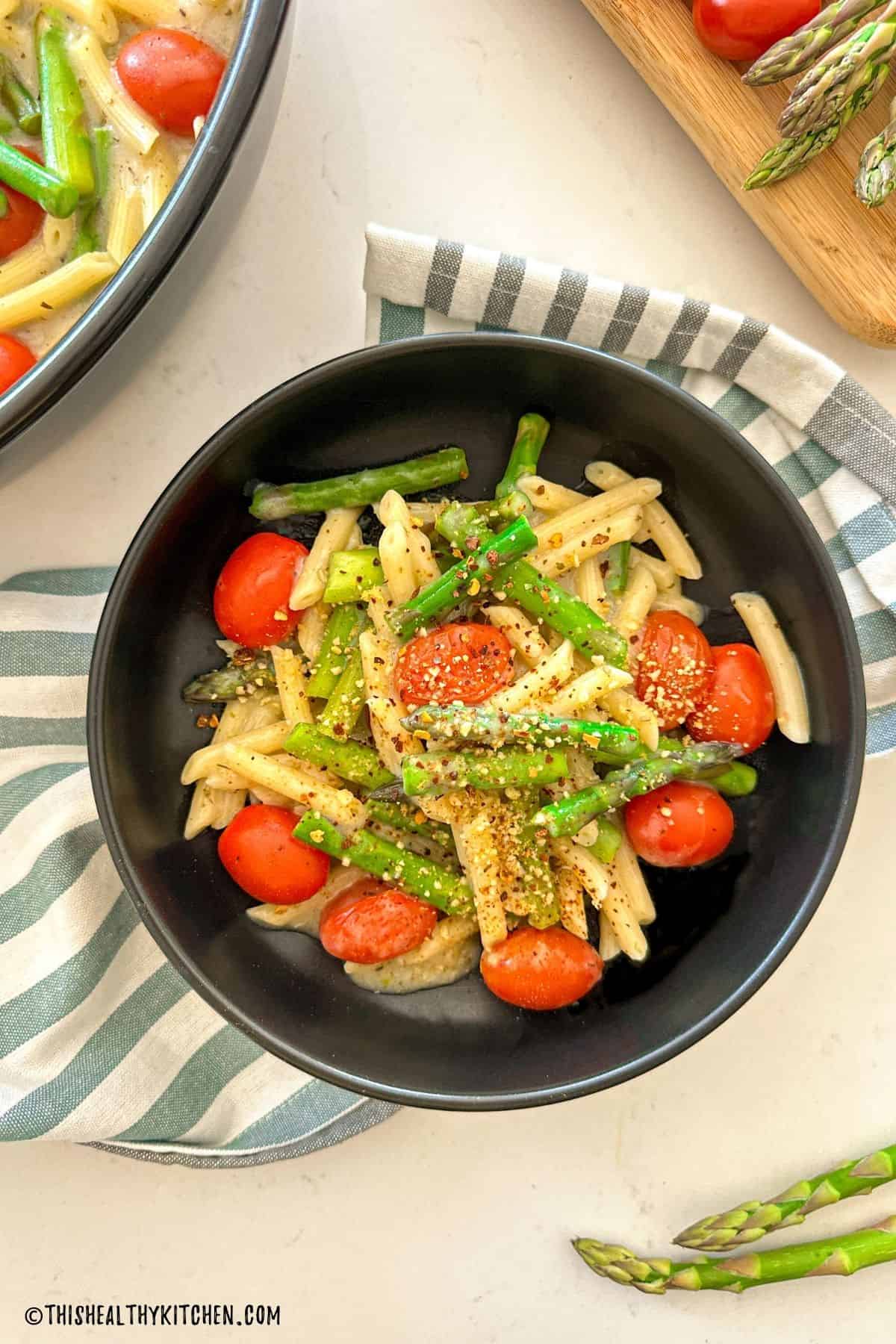 Asparagus and tomato pasta in black bowl with red pepper flakes on top.