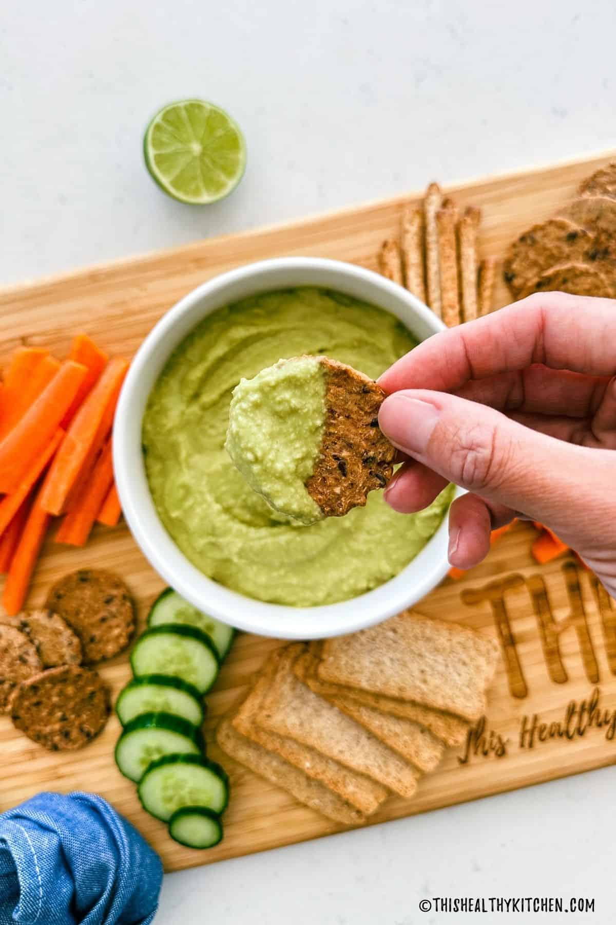Cracker being dipped into bowl of green dip on cutting board.