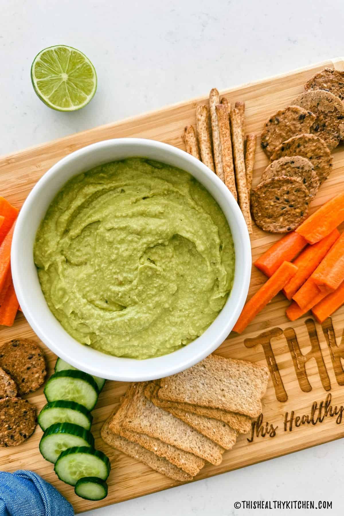 Bowl of jalapeno avocado dip with crackers, carrot sticks and cucumber slices around it.