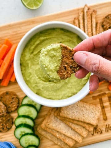 Cracker being dipped into bowl of green dip on cutting board.