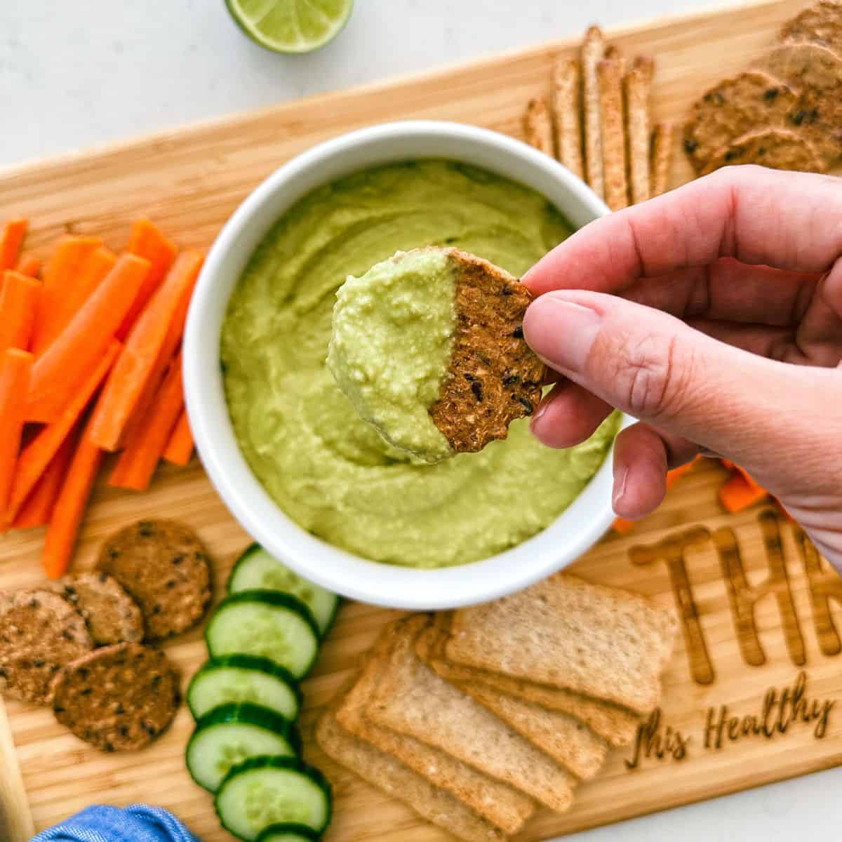 Cracker being dipped into bowl of green dip on cutting board.