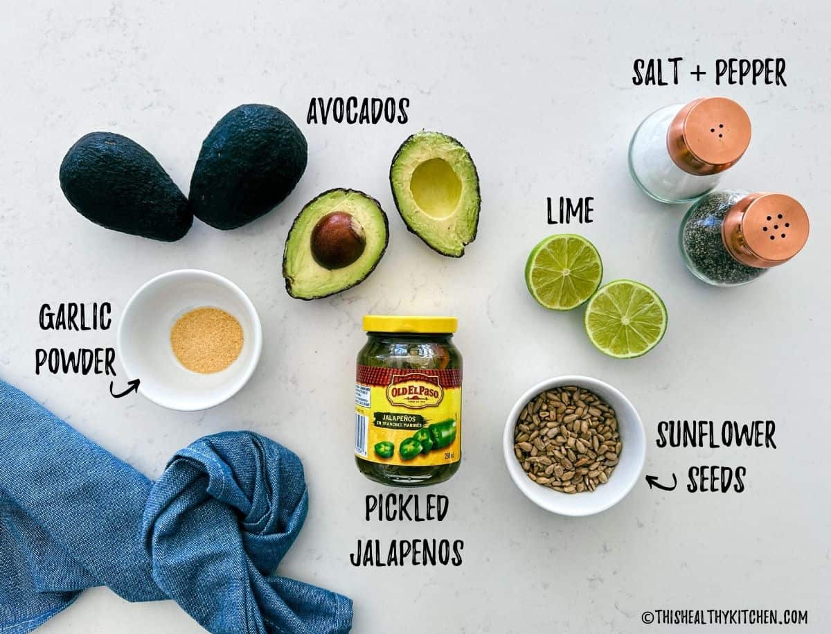 Avocados, limes, jalapenos, sunflower seeds. garlic powder, and salt and pepper on kitchen countertop.