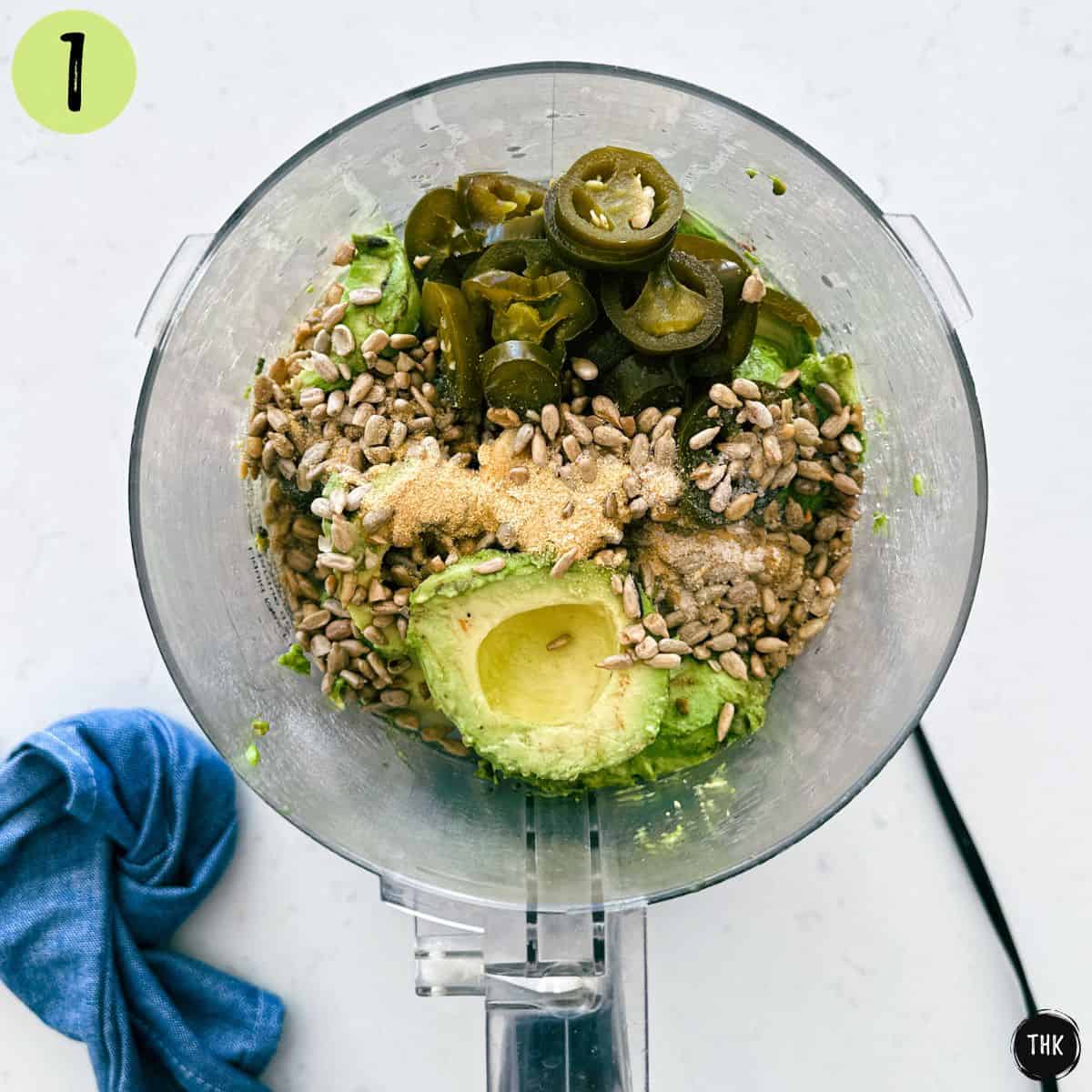 Food processor with jalapenos, avocado, sunflower seeds, and spices inside.