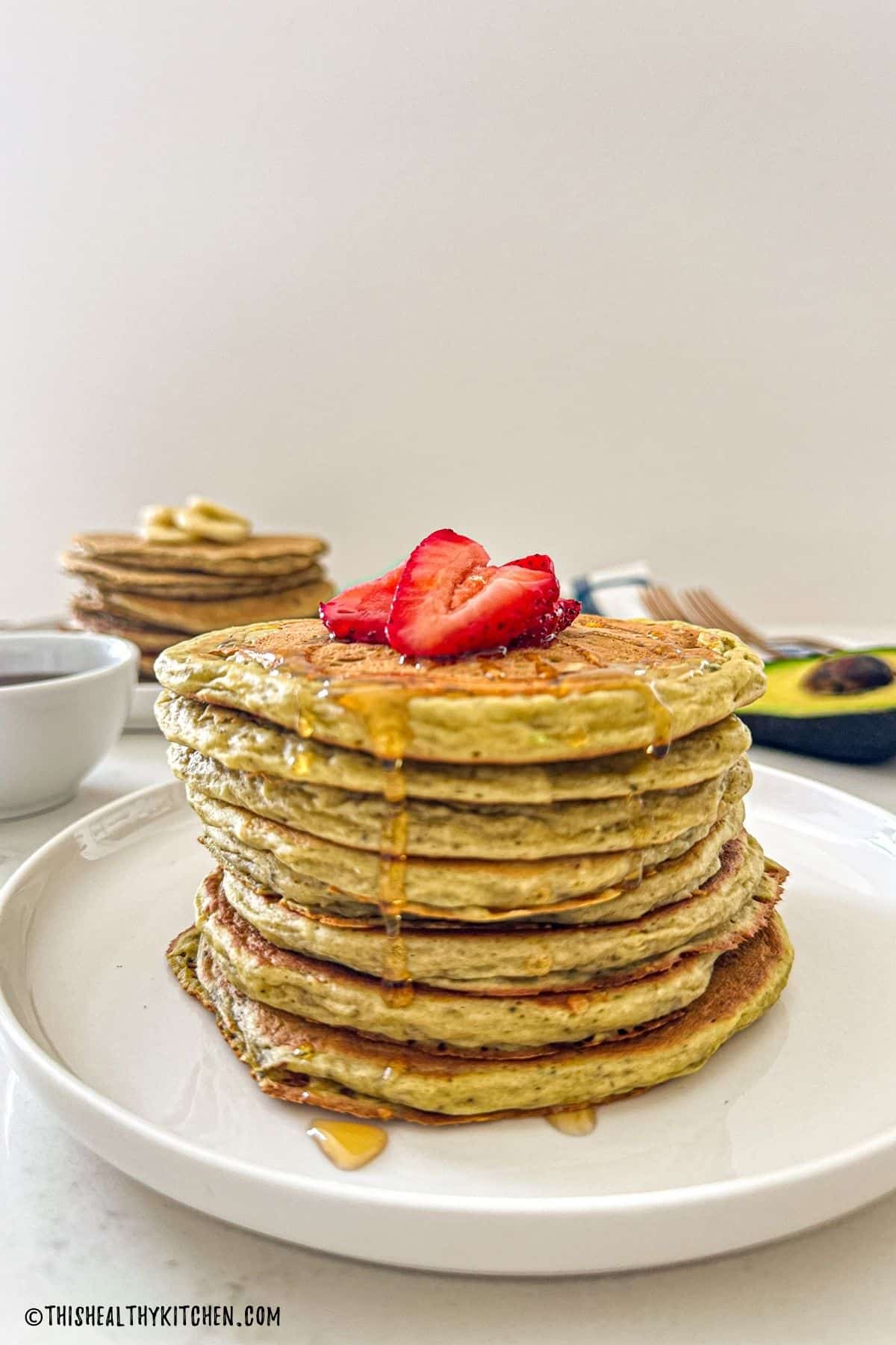 Stack of avocado pancakes on white plate with sliced strawberries on top.