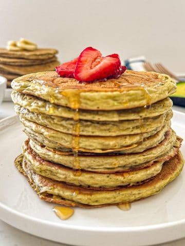 Stack of avocado pancakes on white plate with sliced strawberries on top.