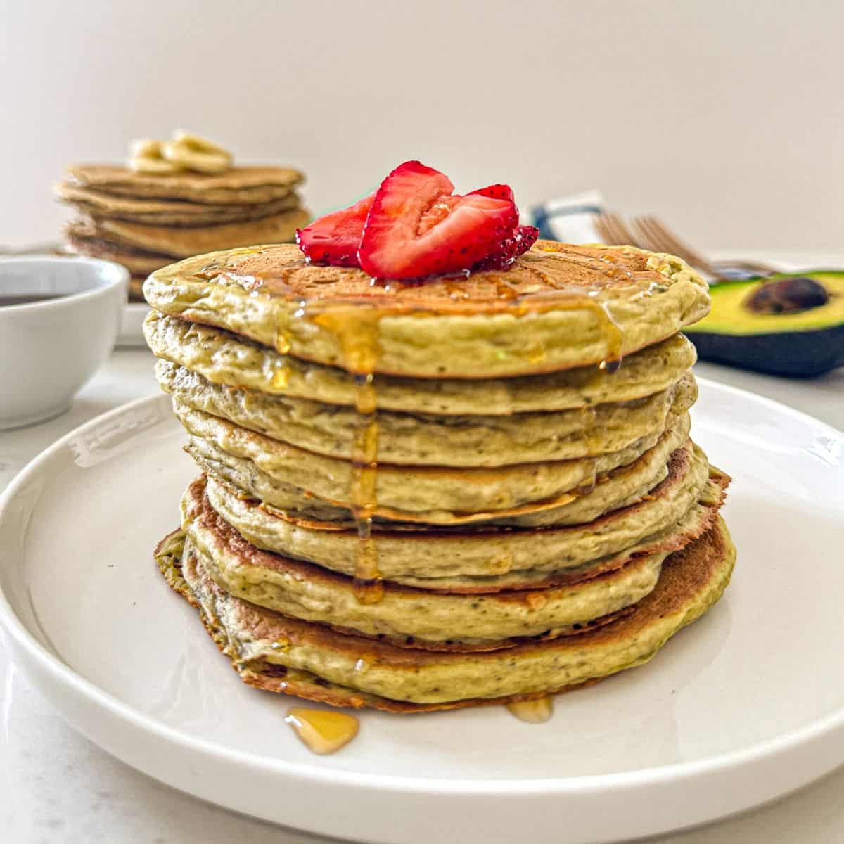 Stack of avocado pancakes on white plate with sliced strawberries on top.