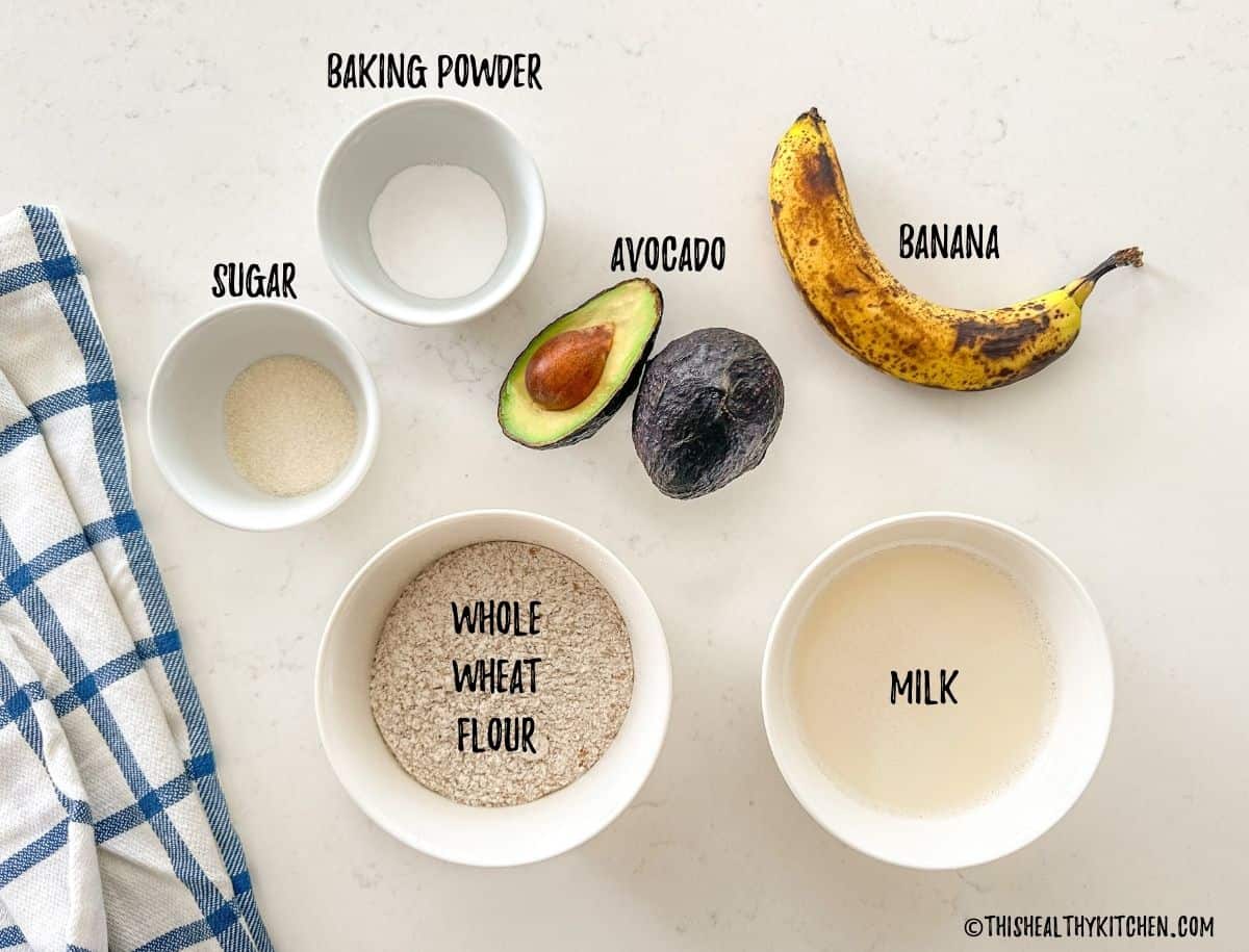 Kitchen countertop with bowls of flour, milk, sugar, baking powder, and a banana and avocado on top.