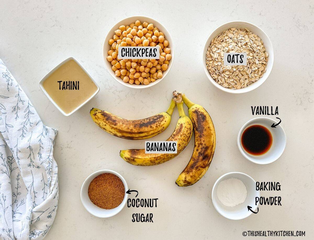 Bananas, bowl of chickpeas, oats, tahini, vanilla, baking powder and sugar on kitchen countertop.