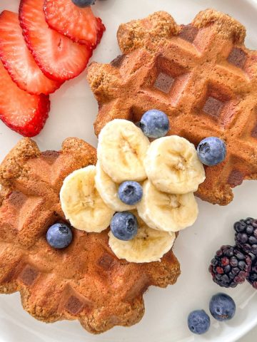 White plate with two banana protein waffles on it, with sliced bananas, strawberries and blueberries on top.