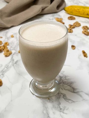 Cup with white smoothie inside and banana and walnuts in background.