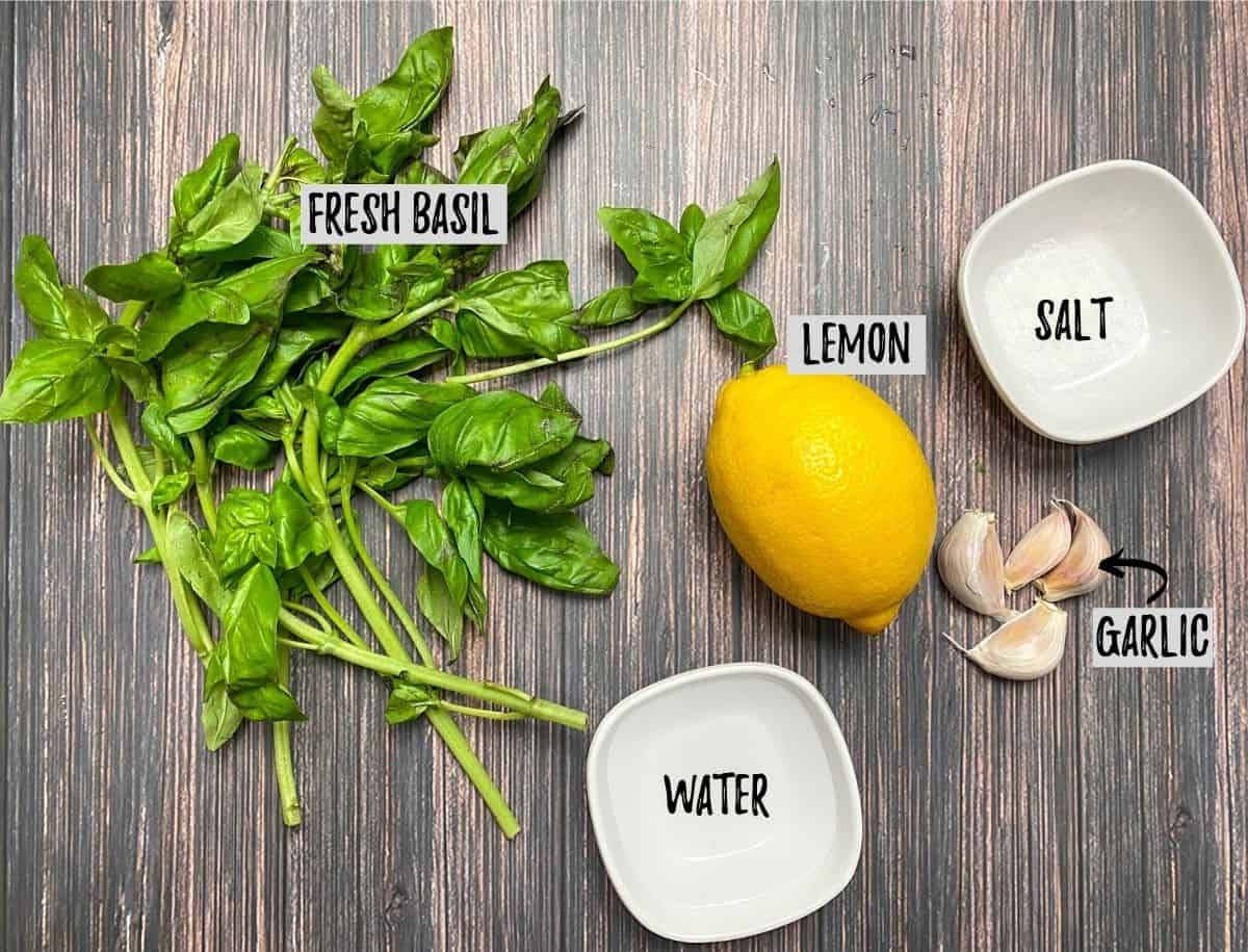 Basil leaves, lemon, garlic cloves and cup of water on brown deck.