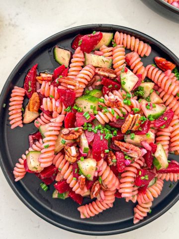 Black plate with rotini pasta, diced beets, cucumber, pecans and chives on top.