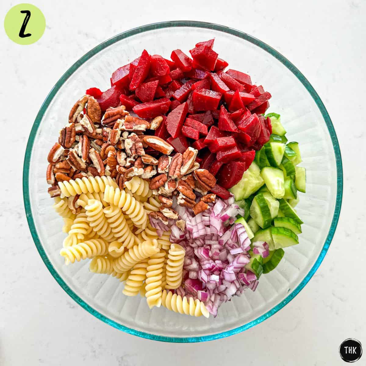 Large glass bowl with rotini pasta, chopped pecans, diced beets, cucumber and red onion.