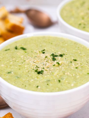 Two bowls of broccoli soup with bread and croutons beside them.