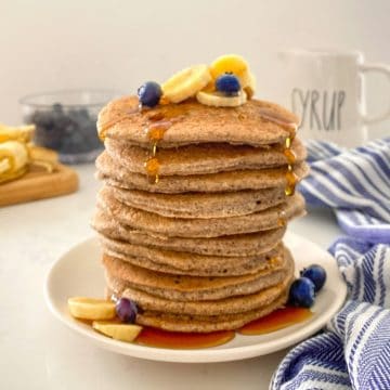 Stack of buckwheat pancakes on plate with banana and blueberries on top.