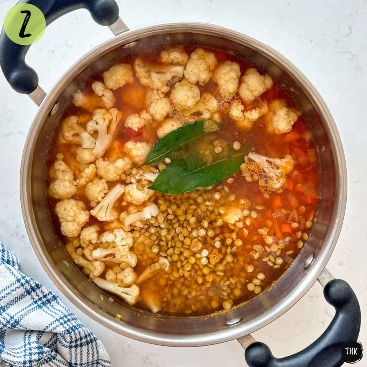 Large pot of soup with cauliflower, lentils, broth, tomato sauce, veggies and bay leaves on top.
