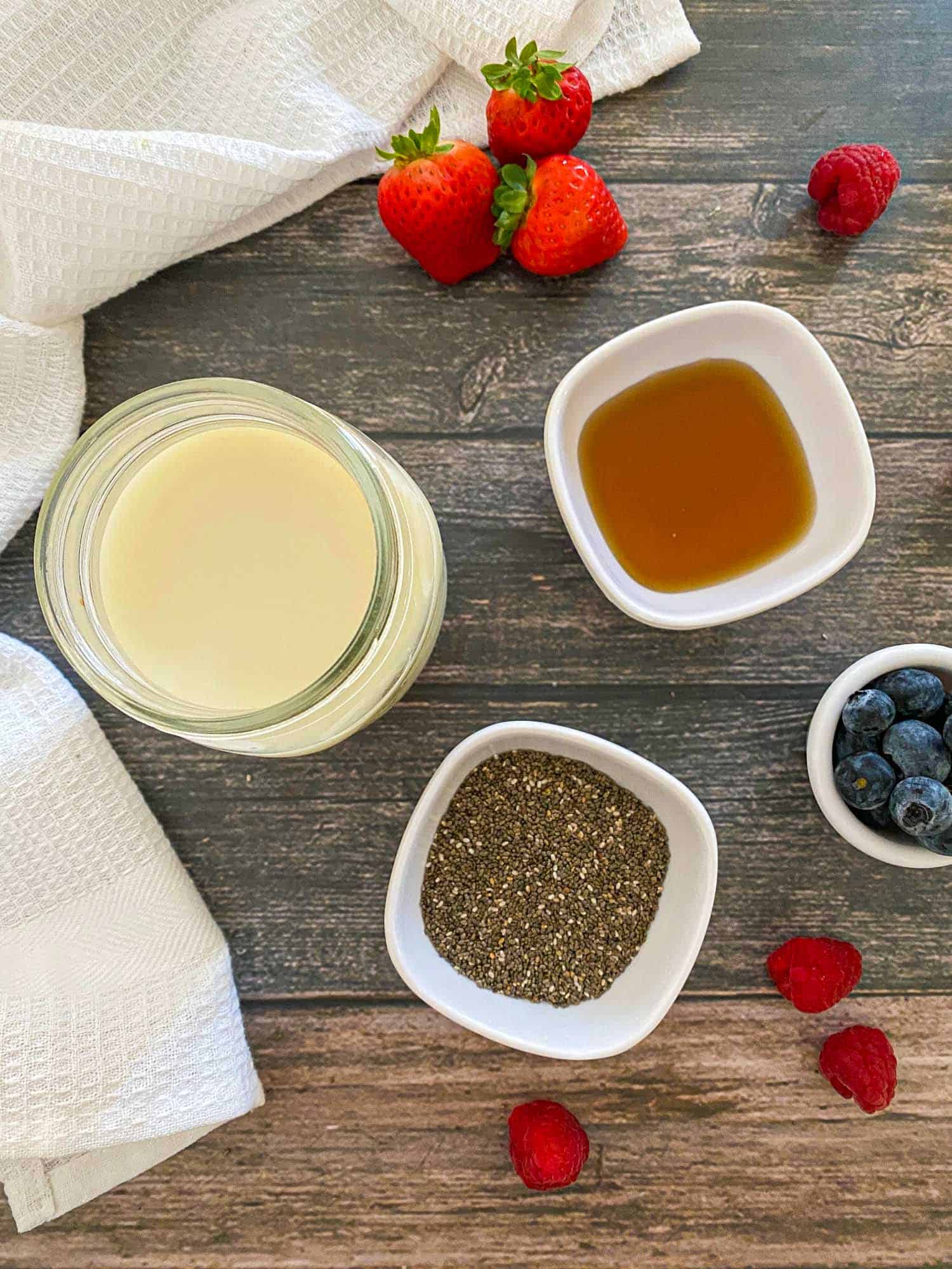 Jar of milk, bowl of chia seeds, bowl of maple syrup and berries on grey deck.