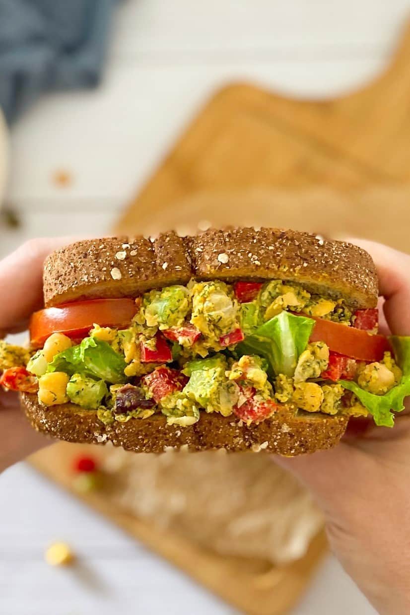 Hand holding up chickpea salad sandwich above cutting board.