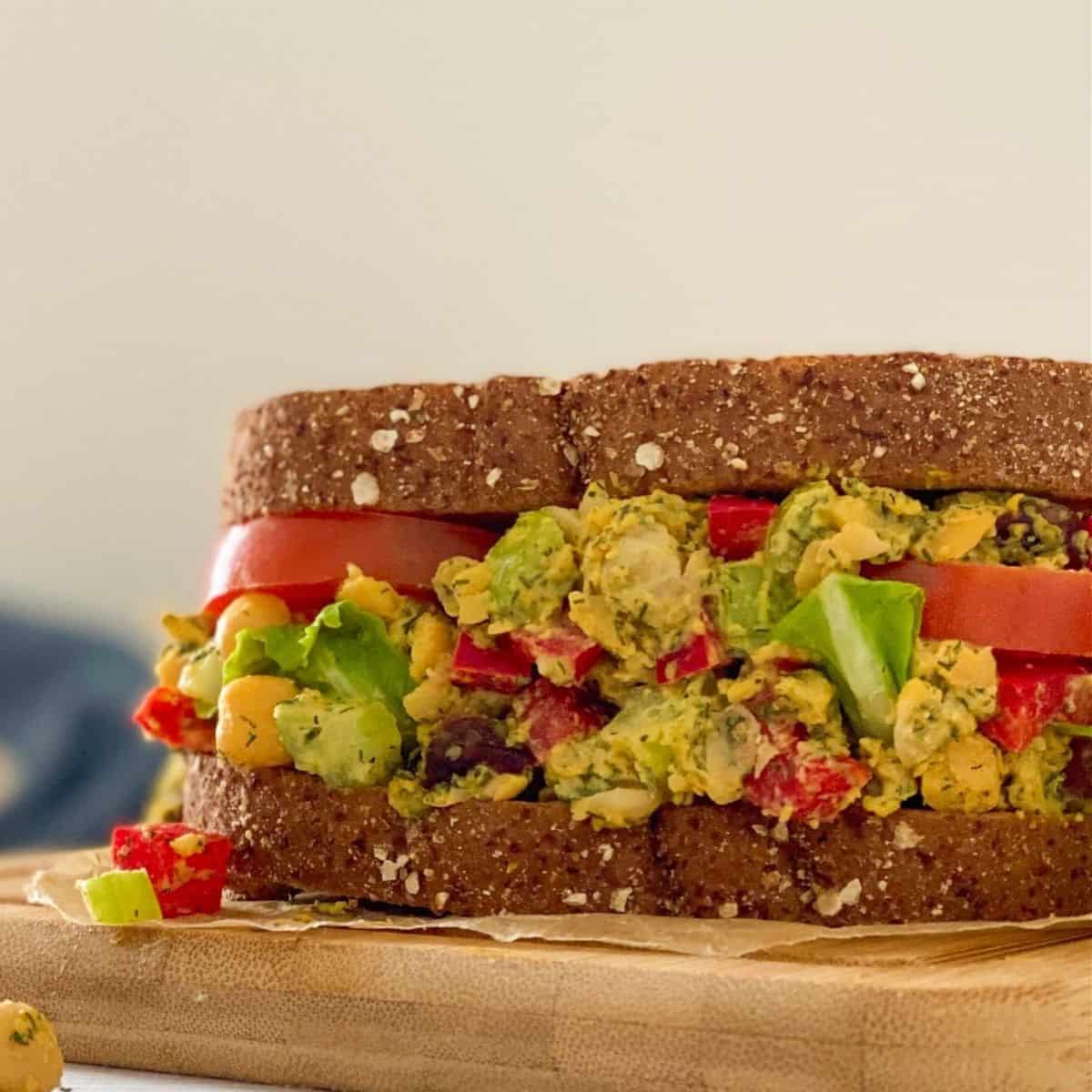 Sandwich resting on cutting board with chickpeas, tomato, celery, peppers and dill.
