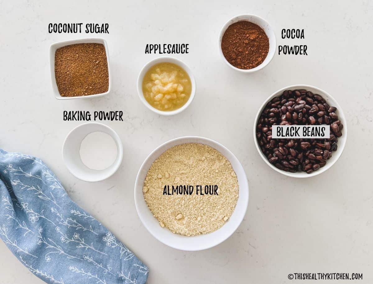 Bowls of almond flour, black beans, cocoa powder, applesauce, sugar, and baking powder on countertop.