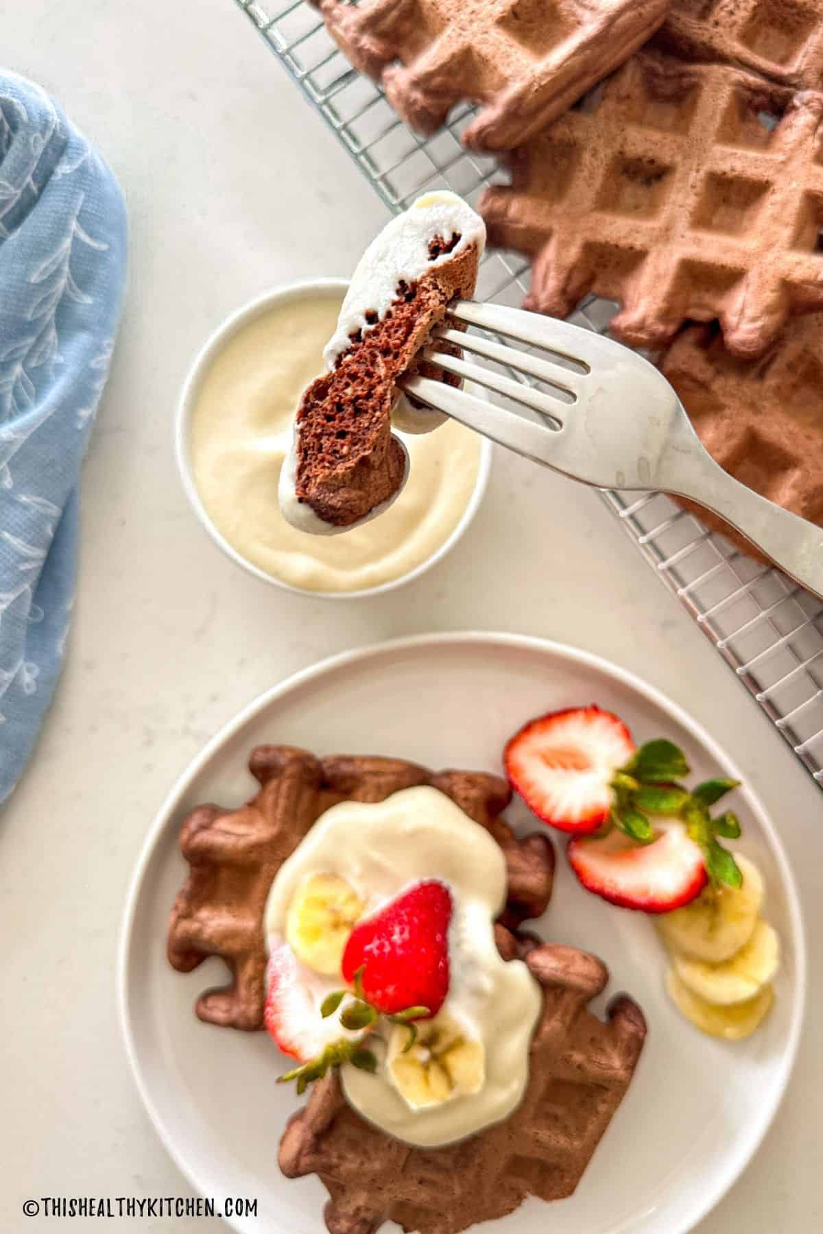 Fork with bite of chocolate waffle on it with more chocolate waffles on plate below.