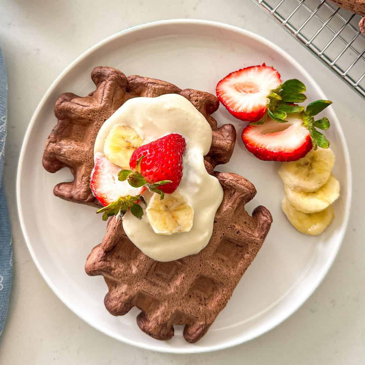 Two chocolate waffles on plate with vegan whipped cream, strawberries and banana on top.