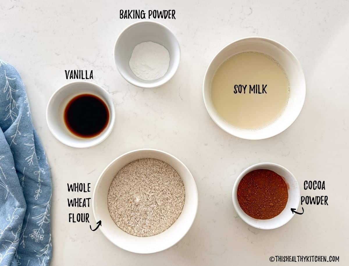 Bowls of flour, cocoa powder, milk, vanilla and baking powder on countertop.