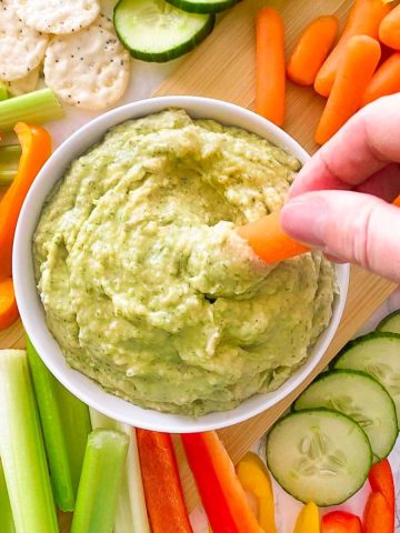 Carrot being dipped into bowl of dill pickle hummus.