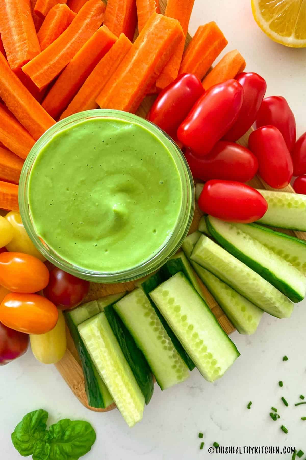 Green dip in glass jar with raw veggies surrounding it.