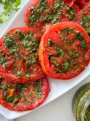 White platter filled with sliced grilled tomatoes with chimichurri on top.