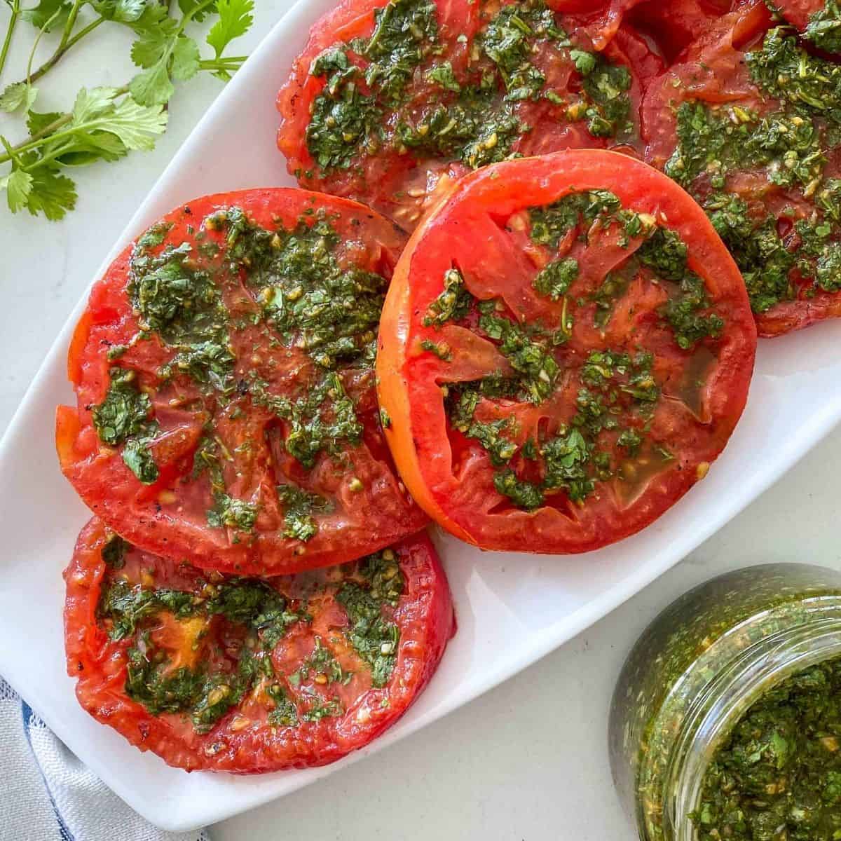 White platter filled with sliced grilled tomatoes with chimichurri on top.