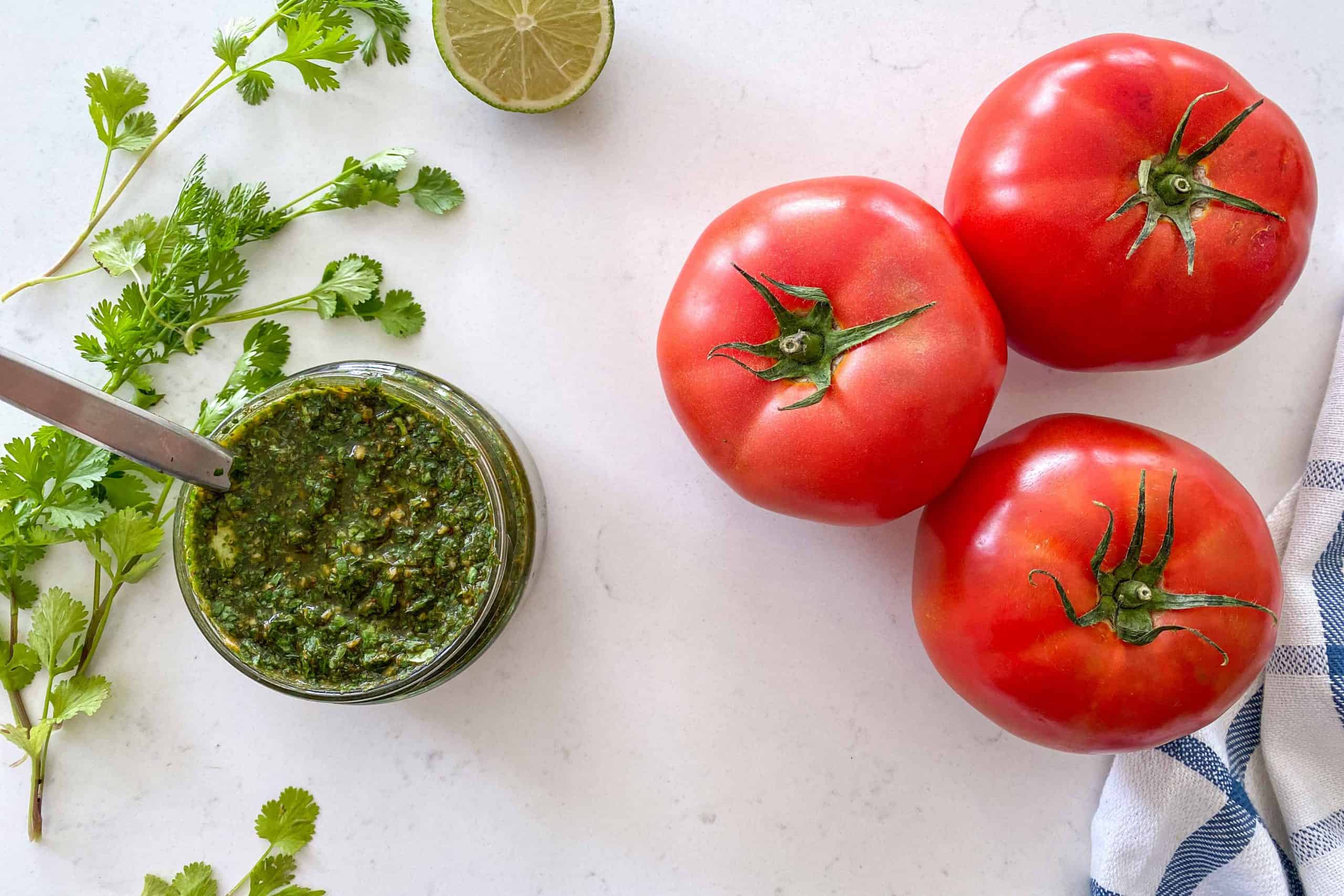 Beefsteak tomatoes on counter top with jar of chimichurri beside them.