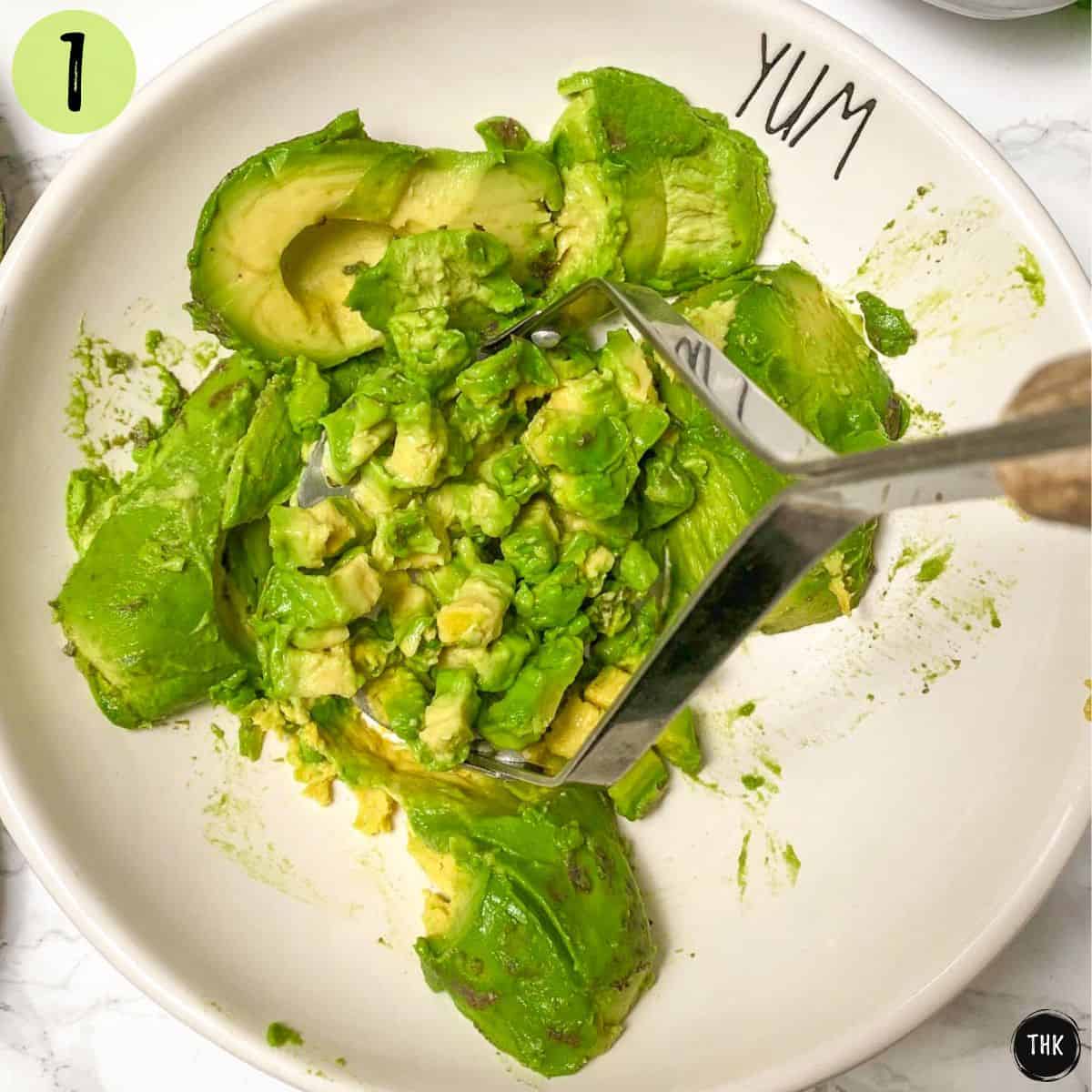 Avocados in white bowl being mashed with potato masher.