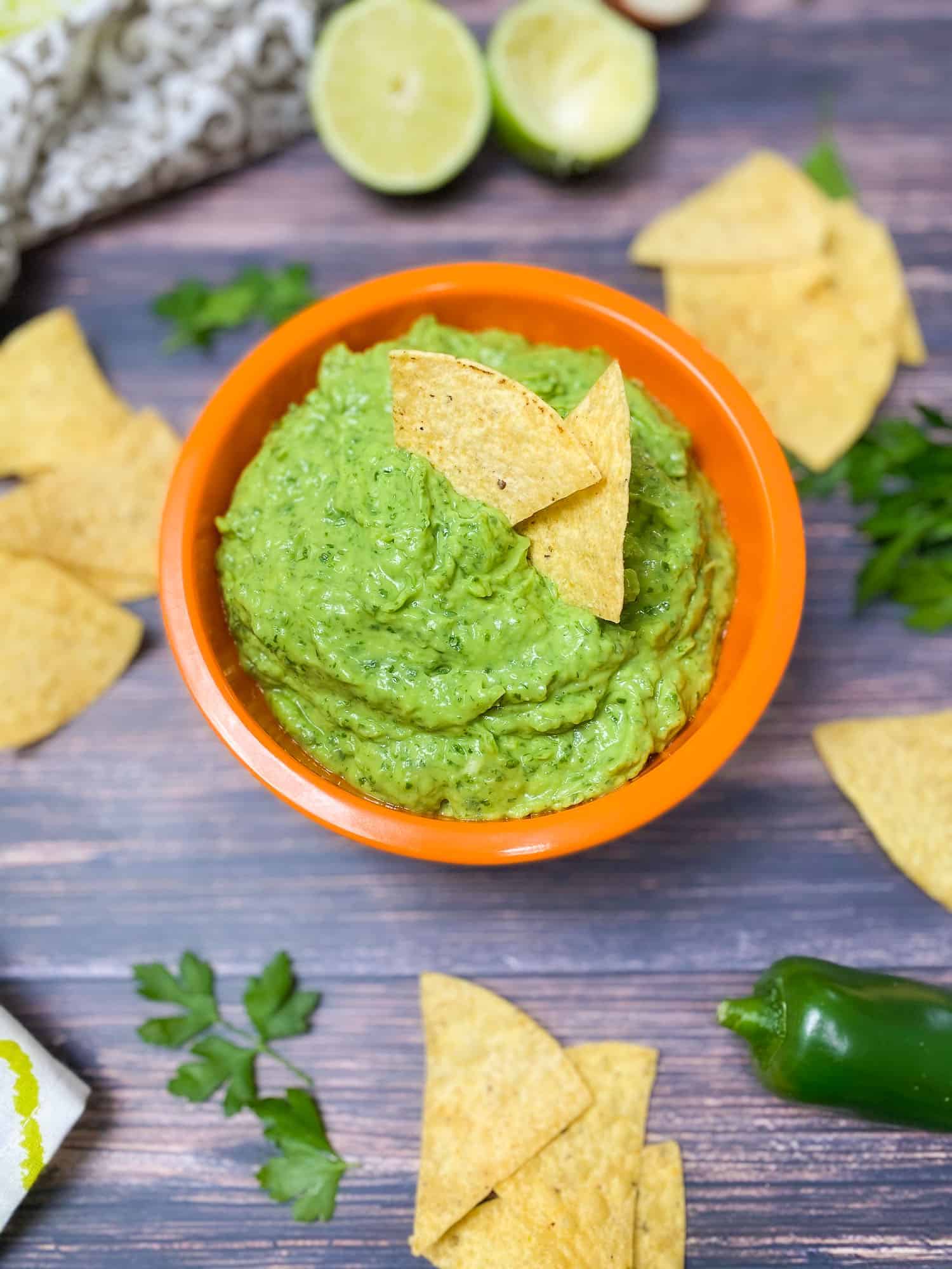 Two chips sitting in bowl of green dip with chips and lime scattered around the bowl.