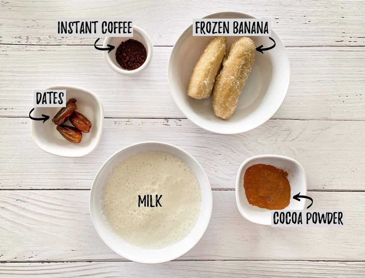 Frozen banana, dates, milk, cocoa powder and instant coffee on white deck.