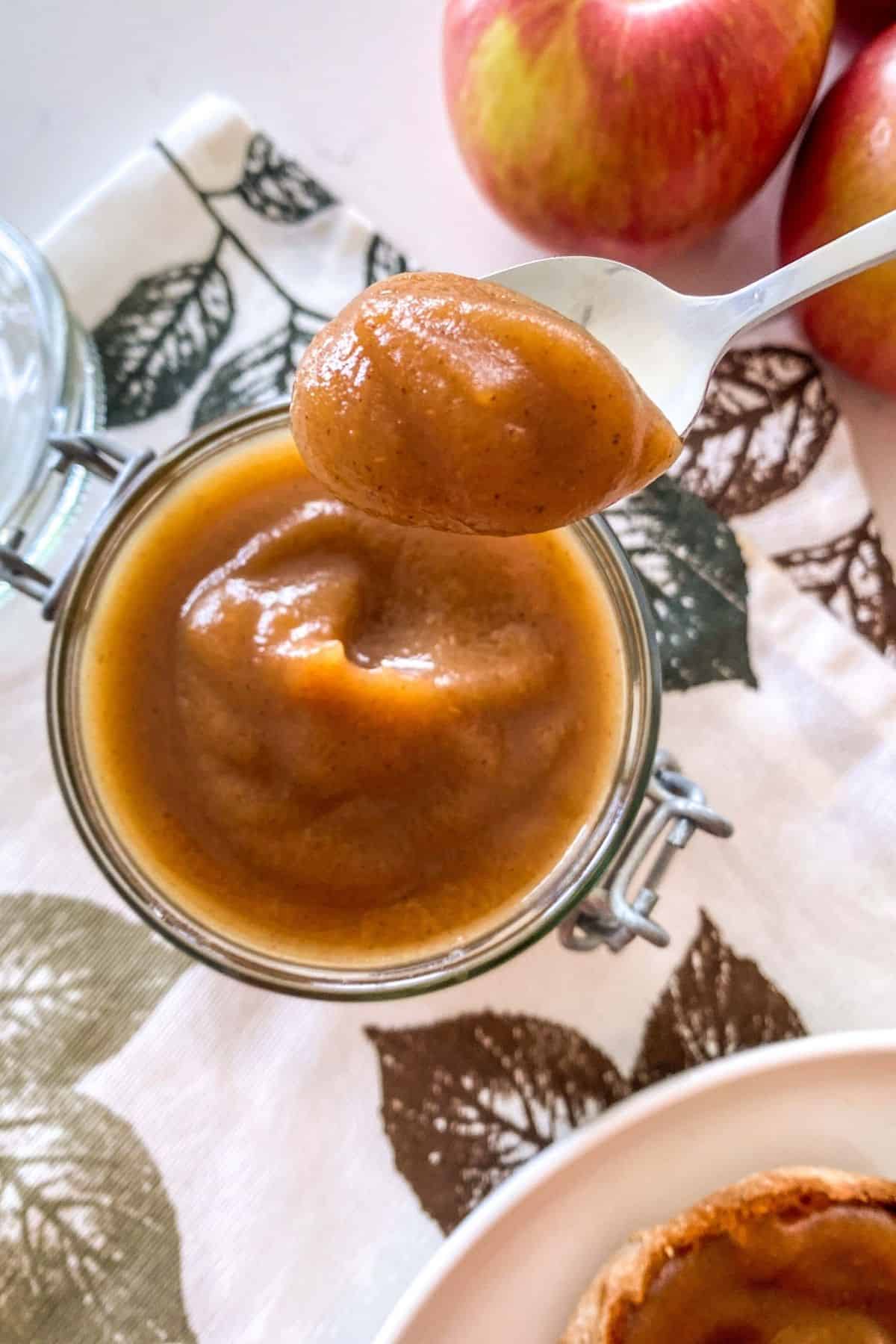 Jar of apple butter with spoonful held up above it and apples in the background.