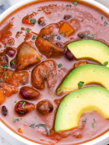 White bowl of tomato bean soup with avocado slices on top.
