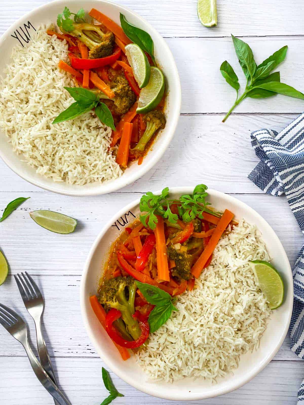 two white bowls with rice, carrots, peppers, broccoli and a red curry sauce with lime wedges and basil garnish