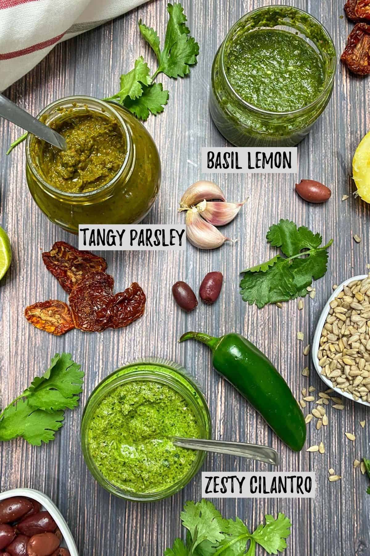 Three glass jars filled with pesto with herbs, garlic and seeds scattered around them.