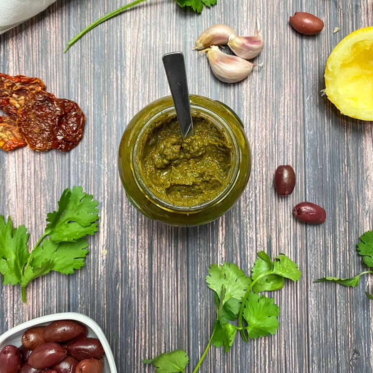 Parsley pesto in glass jar with spoon inside and cilantro, garlic, olives and lemon on the side.