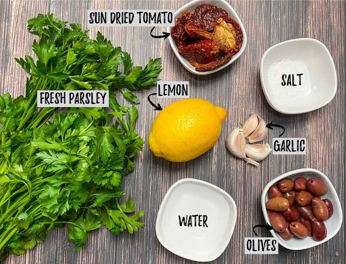 Parsley, lemon, sun dried tomatoes, garlic, olives and bowl of water on brown deck.