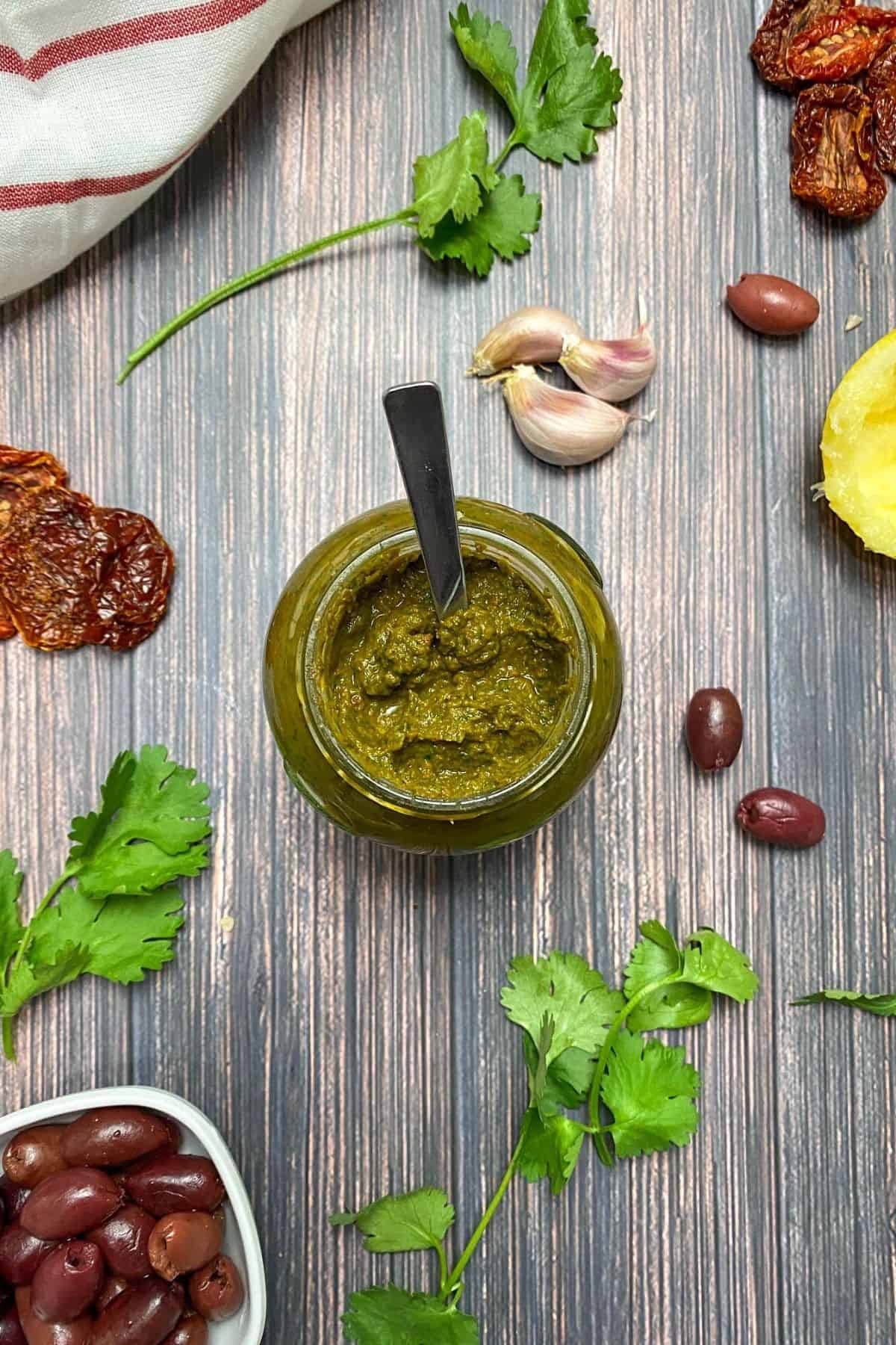 Parsley pesto in glass jar with spoon inside and cilantro, garlic, olives and lemon on the side.